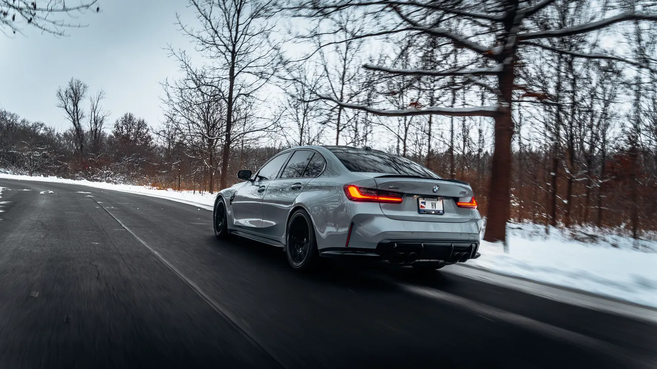 Grey BMW M3 driving on a snowy tree-lined road at dusk