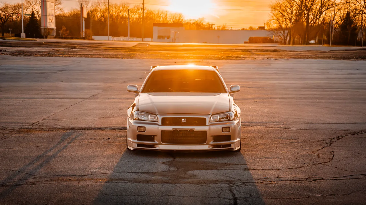 Silver Nissan Skyline R34 GT-R head-on in an empty lot, backlit by a low golden sun