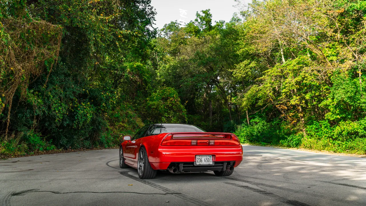 Red Acura NSX shot from the rear three-quarter on a road lined with green foliage